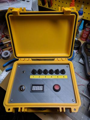 "A photo of a launch controller in a yellow case, with an LED, six black toggle switches, a key switch, a digital meter, and a red launch button."