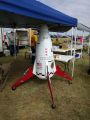 A photo of a white capsule-like rocket on four red lander legs, about six feet tall, under a blue canopy tent.