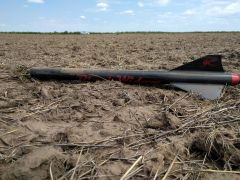 The lower airframe of a black rocket, covered in dust and lying in a dusty field. A silver Volkswagen GTI is in the far distance.